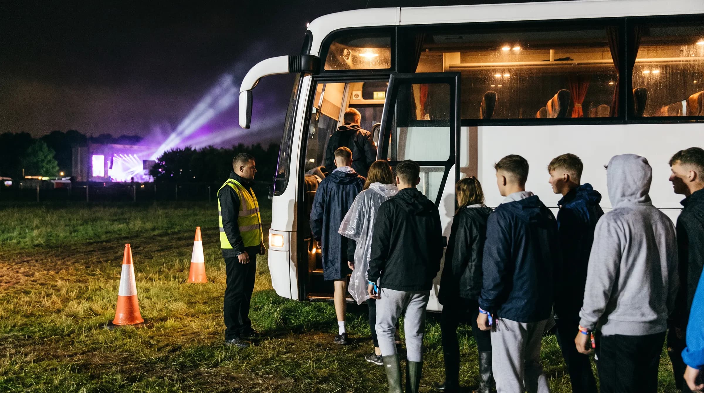 Bezoekers stappen 's nachts in een bus bij de uitgang van een festivalterrein, het podiumlicht nog op de achtergrond