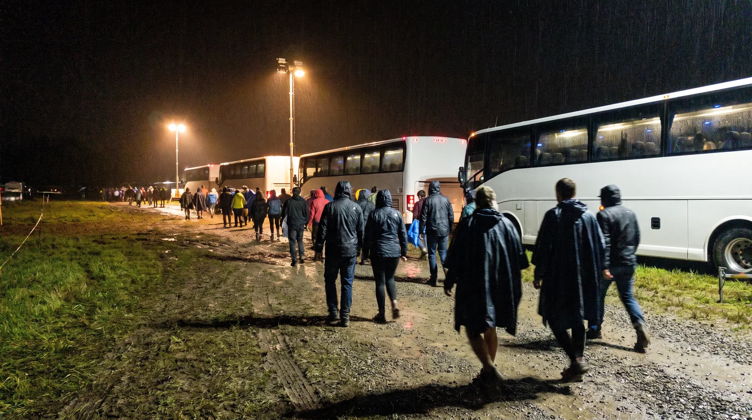 Bezoekers in regenponcho's lopen 's nachts naar de wachtende bussen bij de uitgang van een festival